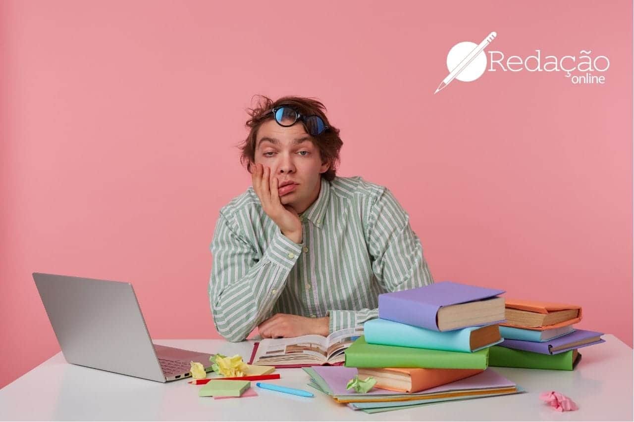 Imagem de um rapaz sentado apoiando a cabeça na mão, com vários livros e um notebook em cima da mesa.