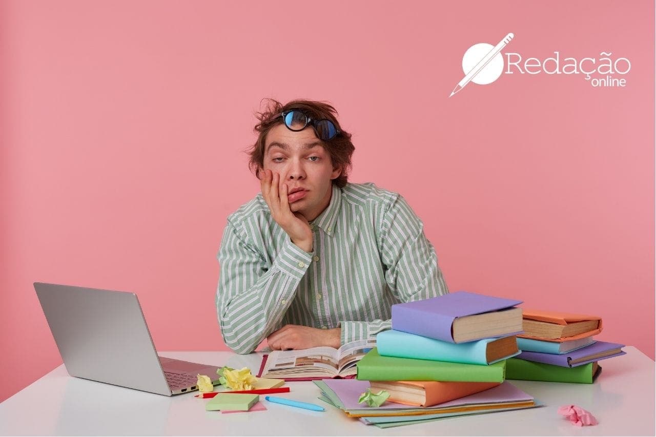 Imagem de um rapaz sentado apoiando a cabeça na mão, com vários livros e um notebook em cima da mesa.